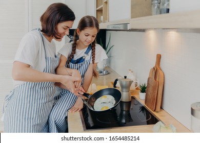 Busy Mother And Daughter Wear Striped Aprons, Pose At Kitchen Near Cooker, Fry Eggs On Pan, Prepare Fast Breakfast, Enjoy Domestic Atmosphere. Mum Teaches Small Kid To Cook. Happy Family Concept