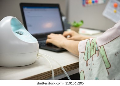 Busy mother or mom pumping breastmilk by Automatic breast pump machine wirh Nursing fabric cover while typing on laptop pc computer on table. Motherhood in corporate office.