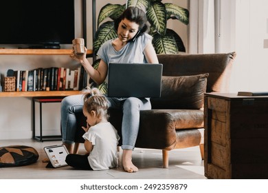 Busy mother multitasks at home, balancing work and childcare, working on a laptop while her daughter plays nearby - Powered by Shutterstock