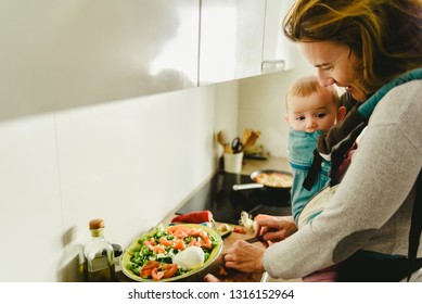 Busy Mother Preparing Food In The Kitchen While Taking Care Of Her Baby, In A Baby Carrier Using The Kangaroo Method.