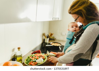 Busy Mother Preparing Food In The Kitchen While Taking Care Of Her Baby, In A Baby Carrier Using The Kangaroo Method.