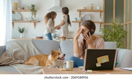 A Busy Mother Trying To Work Remotely With Her Children's At Home. Working From Home During Quarantine Epidemic. On The Background Her Daughters Are Jumping Together Near Lying On The Couch A Cute Dog