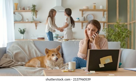 A Busy Mother Trying To Work Remotely With Her Children's At Home. Working From Home During Quarantine Epidemic. On The Background Her Daughters Are Jumping Together Near Lying On The Couch A Cute Dog