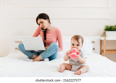 Busy Mother Working On Laptop And Talking On Phone While Her Little Kid Drinking Water From Bottle, Sitting On Bed At Home. Millennial Woman Working At Maternity Leave