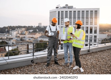 Busy multicultural architects wearing reflective vests and hard hats standing on roof with equipment for quality performance of work. Cooperative people discussing new project in friendly atmosphere. - Powered by Shutterstock