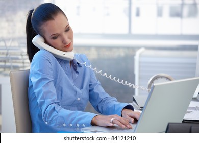 Busy Office Worker Girl Taking Landline Phone Call While Typing On Laptop Computer.?