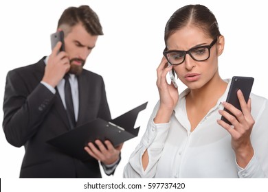 Busy Office Workers Concept. Concentrated Woman Using Two Phones At The Same Time. Unfocused Background With A Man Dressed In Black Suit Having A Conversation.