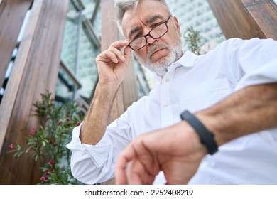 Busy older senior business man looking at wristwatch standing outdoors. Mature old businessman wearing white shirt checking time while waiting or being late for urgent meeting. Time management concept - Powered by Shutterstock