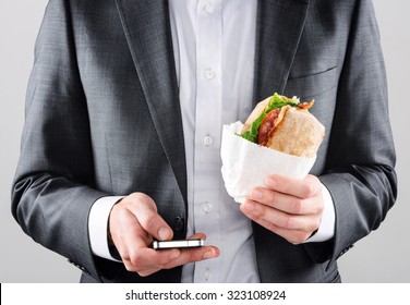 Busy On The Go Business Man In Suit Texting With Smartphone In One Hand And Holding His BLT Ciabatta Roll In The Other