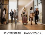 Busy porter pushing cart with baggage while moving among business people interacting on the move in hotel corridor