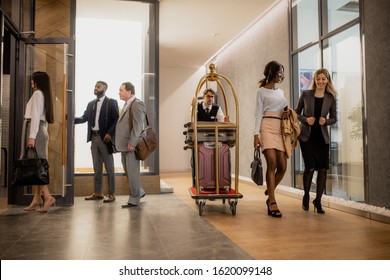 Busy porter pushing cart with baggage while moving among business people interacting on the move in hotel corridor