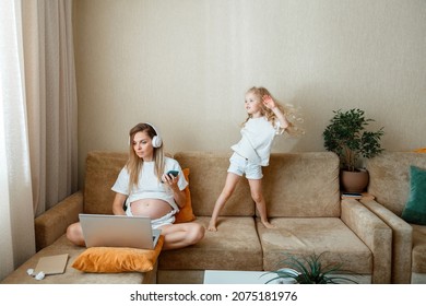 Busy Pregnant Woman Mom Working Remotely Using Laptop Smartphone And Headphones At Home On Couch While Daughter Is Dancing Jumps Having Fun Bouncing Near Distracts Mother. High Quality Photo
