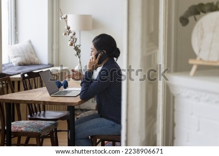 Busy pretty young Indian freelancer woman talking on mobile phone at laptop, discussing job tasks, project on business conversation, using gadgets for communication. Through doorway view, candid shot
