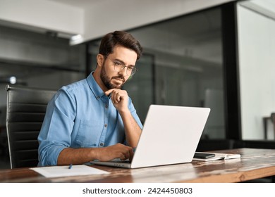 Busy professional business man company employee, young male programmer, latin businessman looking at computer thinking on online investment or software development using laptop working at office.