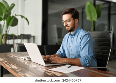 Busy professional business man executive, young male corporate worker, latin businessman wearing glasses typing on computer technology using laptop searching on web working at office workplace desk.