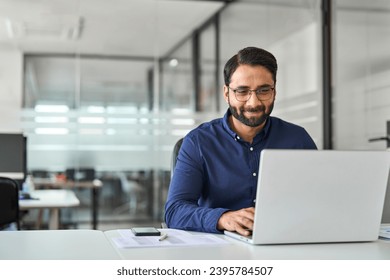 Busy professional Indian business man company employee, male worker software developer, smiling businessman typing on computer technology using laptop working with ai solutions sitting at office desk.