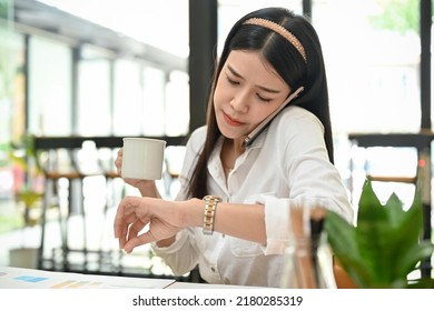 Busy And Professional Millennial Asian Female CEO Or Businesswoman At Her Office Desk, Having A Morning Coffee, Checking Time On Her Wristwatch While Talking On The Phone.