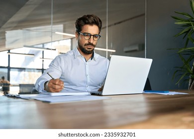 Busy professional young Latin business man company manager executive working in office elearning or having hybrid meeting looking at laptop computer watching webinar writing notes sitting at desk. - Powered by Shutterstock