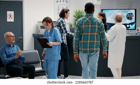 Busy Reception Desk With Many Patients Waiting To Attend Consultation, Trying To Write Medical Report In Hospital Lobby. Diverse People Having Appointments In Waiting Room Area.