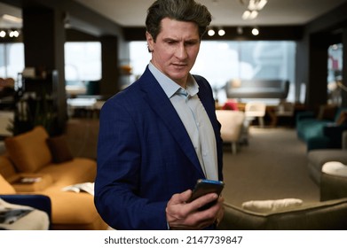 Busy Retail Assistant, Successful Sales Manager, Confident Business Owner, Entrepreneur, Handsome Imposant Caucasian Man In Business Suit Holds Mobile Phone In The Furniture Store Showroom