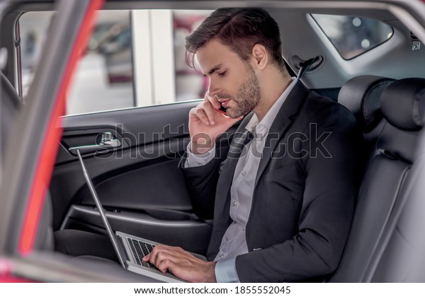 Busy schedule. Brown-haired male sitting\
at backseat of car with laptop, talking on\
phone