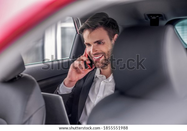 Busy schedule. Smiling male sitting at\
backseat of car with laptop, talking on\
phone
