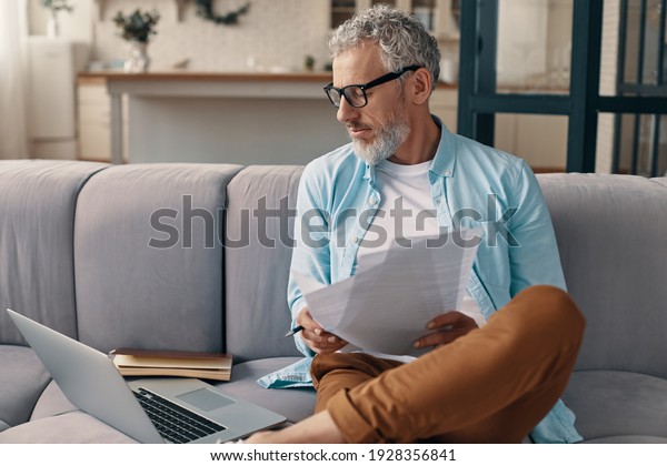 Busy senior man checking the papers and\
using laptop while sitting on the sofa at\
home