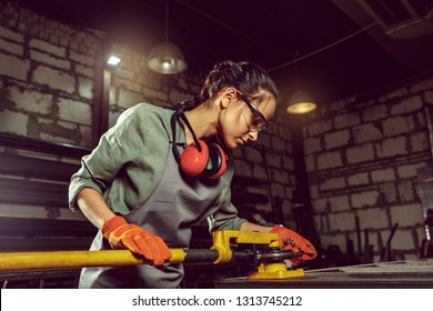 Busy And Serious Craftswoman Grinding Timbers With Special Machine. Beautiful Woman Wearing Safety Glasses. Concept Of Joiner's Shop And Woodworking. Gender Equality. Male Profession