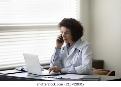 Busy serious mature female chief doctor speaking on mobile phone in office, using laptop computer, giving consultation to patient, checking medical records on online app. Medical career, communication
