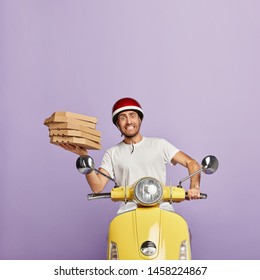 Busy service worker overload with many cardboard boxes of pizza, cannot drive motorbike well, has puzzled look, wears helmet, tries to deliver fast food in time. Multitasking. Delivery concept