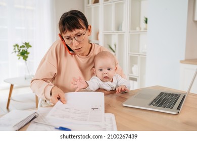 Busy Single Mother Taking Phone Call And Trying To Solve Family Promlems While Taking Care Of Her Little Baby. Multitasking Stressed Mom Talking On Cellpphone, Sitting At Desk, Holding Her Toddler