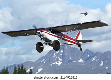 Busy Skies Over Anchorage, Alaska.  A Super Cub Bush Plane Departs The Lake Hood Gravel Runway, While Another One Circles To Land Behind.