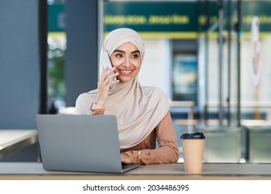 Busy Smiling Attractive Young Arab Lady In Hijab Calls On Phone, Sits With Laptop And Coffee In Cafe Outdoor. Freelancer Female Working While Sitting In City, Digital Nomad, Independent Contractor