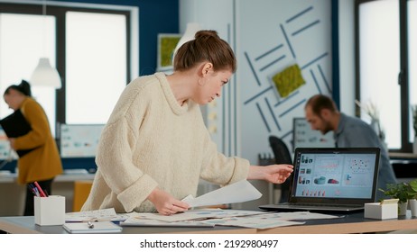 Busy Startup Employee Looking At Papers With Business Data Comparing Numbers With Laptop Screen Standing At Desk. Woman Analyzing Profit Indicators Between Pie Charts And Sales Data.