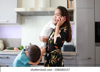 Busy And Stressed Mother Talking On Phone With One Hand Carrying Her Little Infant Baby And Bother Boy Crying And Annoying On Kitchen Background. Housewife Overwork