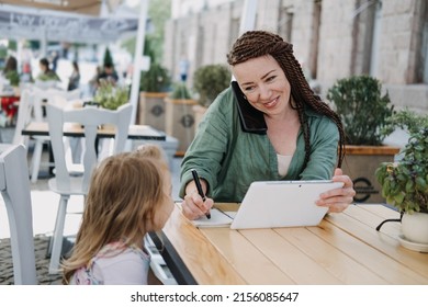 Busy Successful Mother And Business Owner With Cell Phone And Tablet Having Breakfast With Her Little Baby Daughter In Street Cafe In The Summer Day