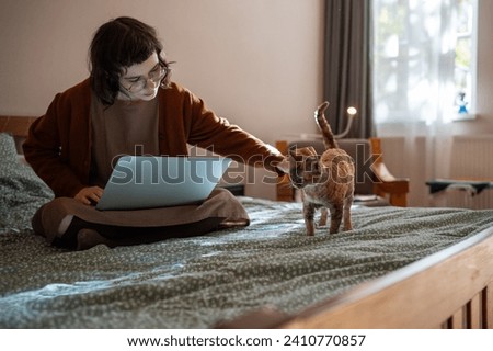 Busy teenager pet owner sitting on bed, working on laptop computer as freelancer. Beloved purring ginger breed cat Devon Rex diverting girl attention, requiring to stroke, caress, amuse, play