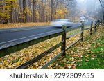 Busy traffic on curved country road with blurry moving cars along weathered wooden fence and colorful autumn leaves at early foggy morning in Rochester, New York, USA. Roadside autumn landscape