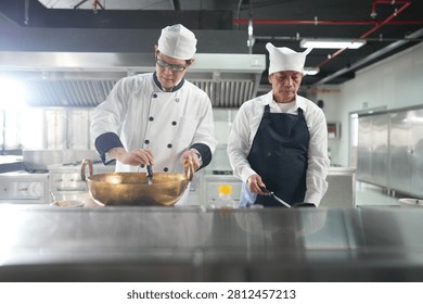 In a busy training kitchen an experienced head chef in a white toque lifts the ladle from the brass wok in the pro kitchen during a practical vocational lesson.