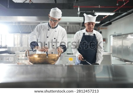 In a busy training kitchen an experienced head chef in a white toque lifts the ladle from the brass wok in the pro kitchen during a practical vocational lesson.