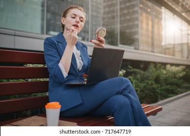 Busy Woman Is In A Hurry, She Does Not Have Time, She Is Going To Do Make Up And To Work On The Laptop . Worker Applying Lipstick In The Street And Working On PC At The Same Time.  