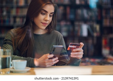 Busy Woman Sitting In A Cafe With Credit Card And Smartphone While While Paying Bills Online.Woman Pays For Purchases With A Card And Phone. Makes Shopping In Popular Web Store.