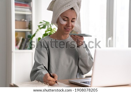 Busy woman with towel on head and patches under eyes, working from home on laptop during lockdown, records a voice message on smart phone, makes notes. Face skin care beauty. Real life. 