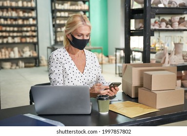 Busy at work. Attractive mature woman, small business owner wearing protective mask using her smartphone while sitting at the desk in her craft pottery shop - Powered by Shutterstock