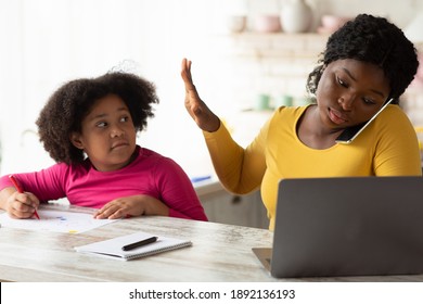 Busy Working African American Mother Ignoring Her Little Daughter Asking For Attention While Drawing In Kitchen. Young Woman Showing Not Now Gesture With Palm, Talking On Cellphone And Using Laptop