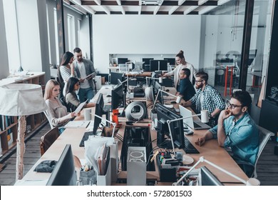 Busy Working Day In Office. Group Of Young Business People In Smart Casual Wear Working And Communicating While Sitting At The Large Desk In The Office Together