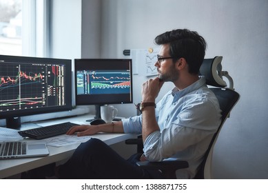 Busy working day. Side view of successful trader or businessman in formal wear and eyeglasses working with charts and market reports on computer screens in his modern office
