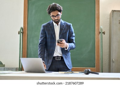 Busy young adult indian business man manager using cell phone apps working on laptop standing in office at work. Arab teacher or coach using technology devices for business and teaching in classroom