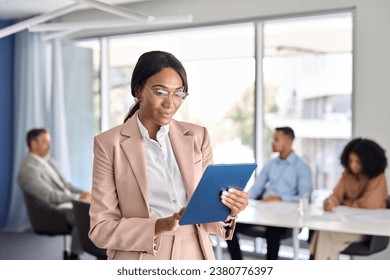 Busy young African American business woman using tab standing at office meeting. Smiling confident professional company executive manager working on digital tablet tech software in board room. - Powered by Shutterstock