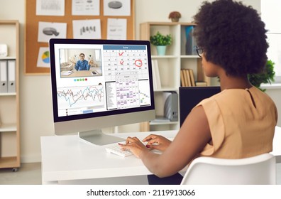 Busy young Afro American woman working at office desk with desktop computer, chatting with coworker, organizing schedule in calendar planner, having conference call with colleague. View over shoulder - Shutterstock ID 2119913066